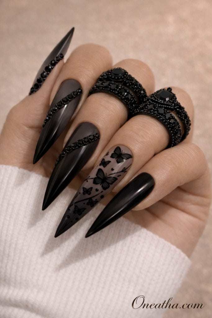 Matte black stiletto nails with butterfly designs and subtle rhinestone accents on a hand with black statement rings and a neutral background. Elegant black butterfly nail design with soft matte finish and luxe detailing for a bold, modern manicure look.