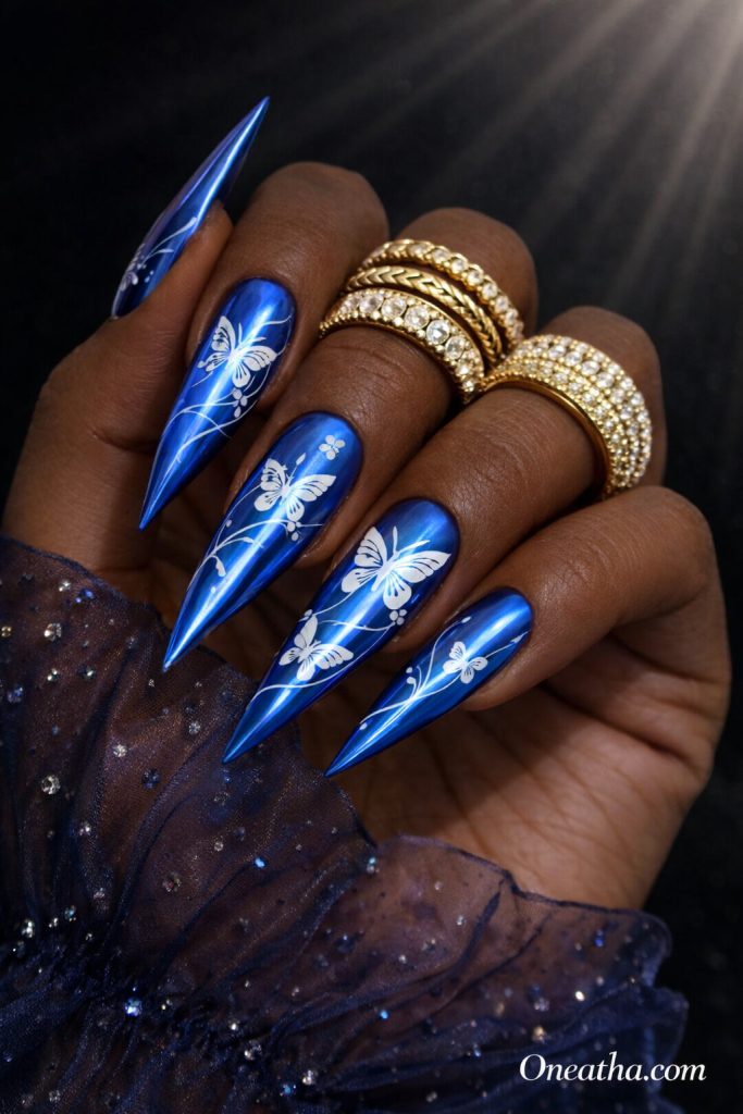Deep cocoa-toned hand with long stiletto blue chrome nails featuring white butterfly designs and flowing blue line art. The hand is styled with chunky gold rings and a sheer navy embellished blouse against a dark luxury background with soft light beams.
