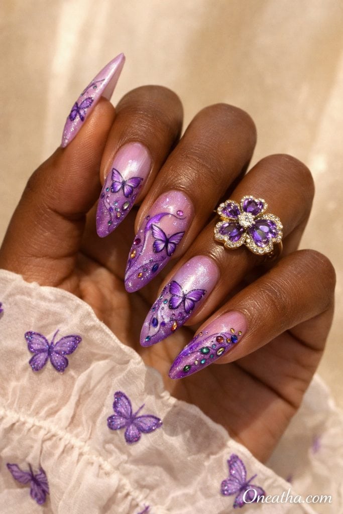 Purple butterfly nails with pink ombre base, glossy stiletto nails featuring butterfly nail art, shimmer glitter, and colorful rhinestone accents on a warm-toned hand with a statement ring