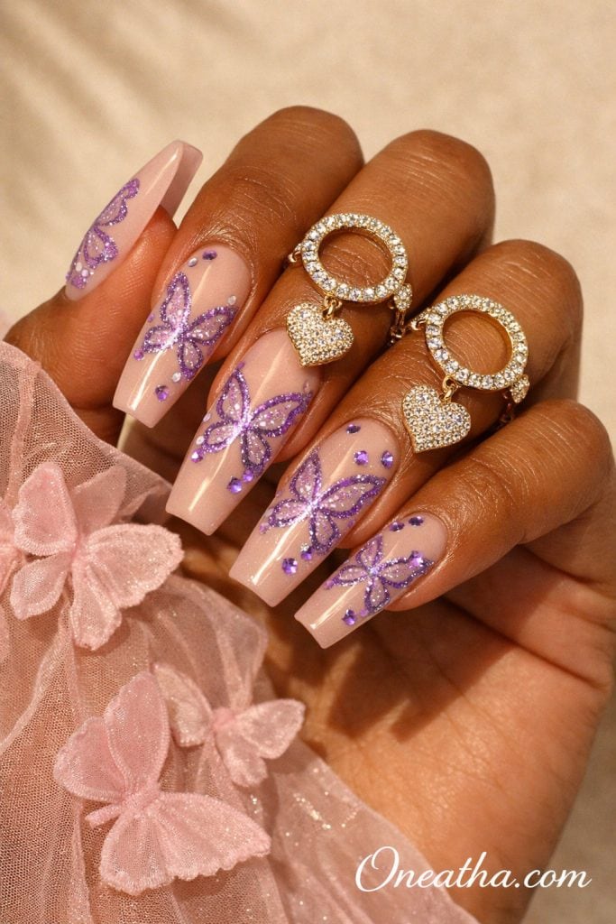 Nude pink and purple butterfly nails with glitter butterfly designs, rhinestone accents, and glossy square acrylic nails, soft glam luxury manicure on warm-toned hand with gold rings, feminine nail inspiration