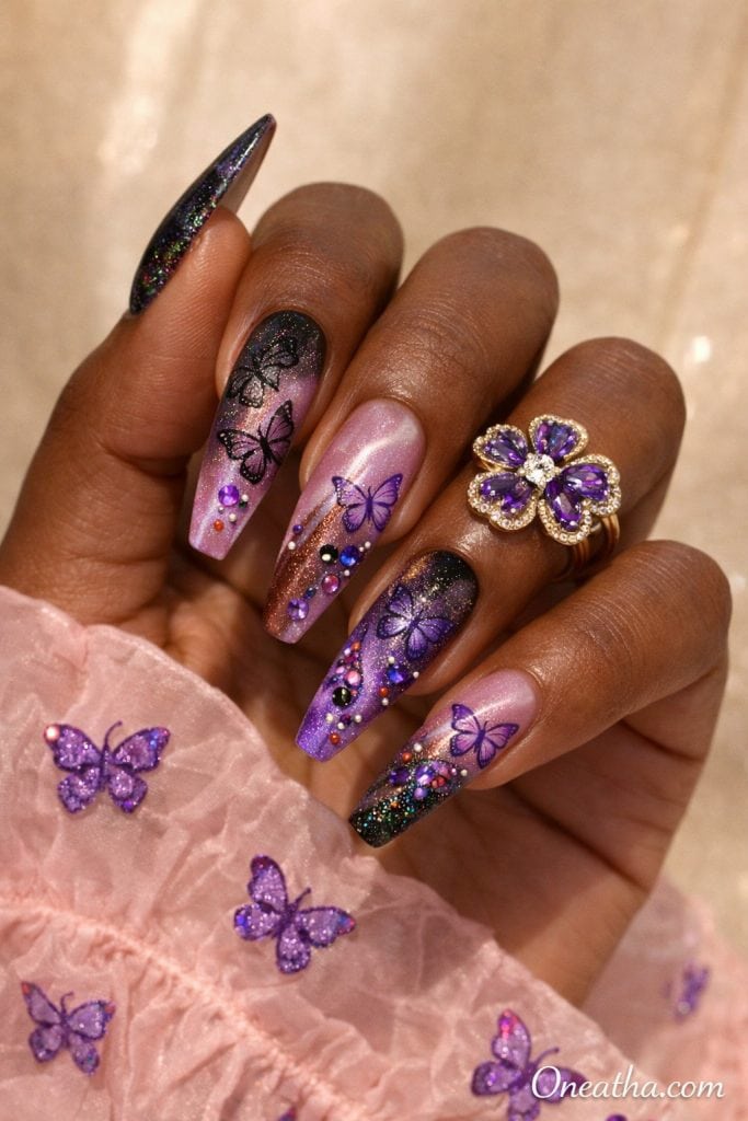 Purple butterfly nails with pink ombre base, black and purple glitter tips, rhinestone accents, and glossy stiletto acrylic nails featuring butterfly nail art on a warm-toned hand with gold ring, luxury nail inspiration