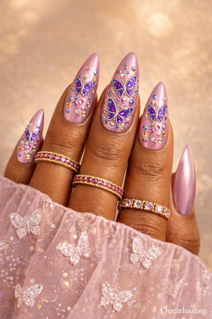 Pink and purple butterfly nails with chrome pink base, rhinestone accents, glossy stiletto acrylic nails featuring butterfly nail art, luxury nail inspiration on warm-toned hand with gold rings