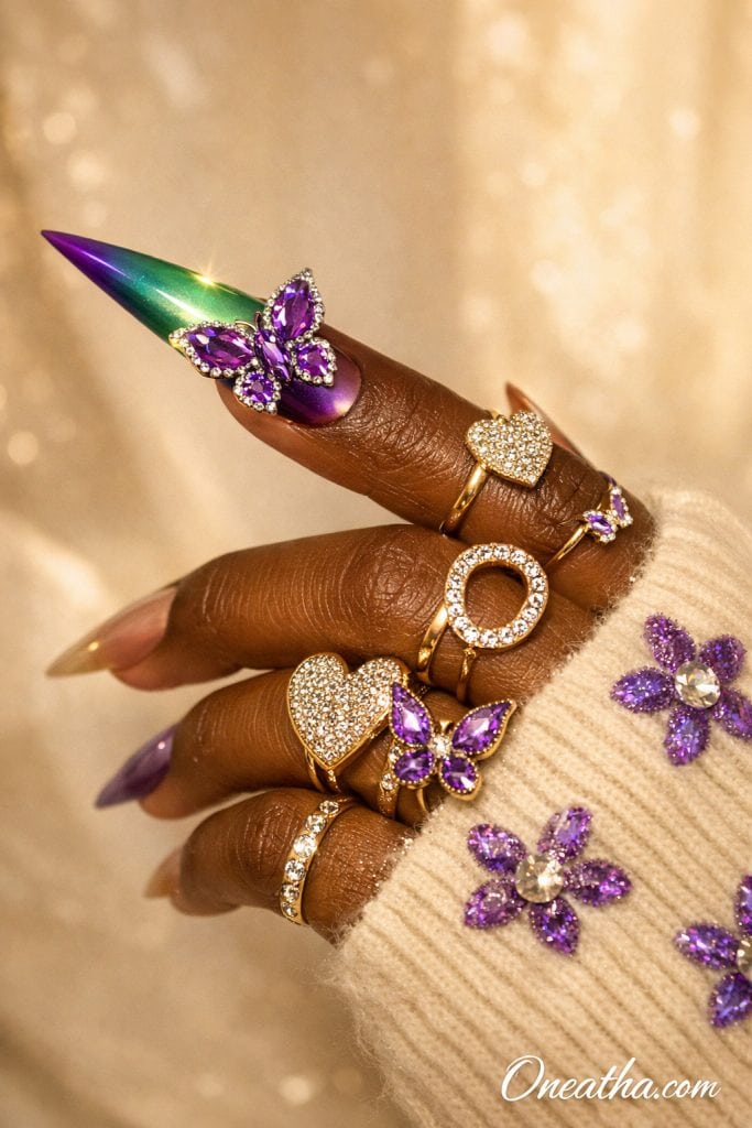 Purple butterfly nails with chrome color shift finish, green and purple ombre stiletto acrylic nails with 3D gemstone butterfly design and rhinestone accents, luxury nail inspiration on warm-toned hand with gold rings