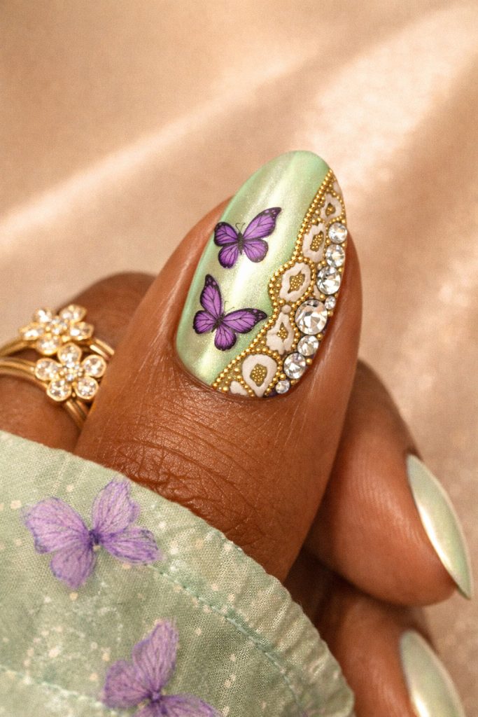 Purple butterfly nails with green chrome base, gold detailing, rhinestone accents, glossy almond acrylic nails, delicate butterfly nail art design on warm-toned hand with gold rings, luxury nail inspiration