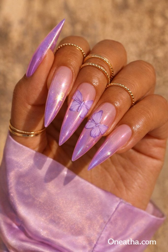 Purple butterfly nails with lilac chrome shimmer, soft pink nude base, glossy stiletto acrylic nails, butterfly nail art design, iridescent purple nails on warm-toned hand with gold rings, luxury nail inspiration