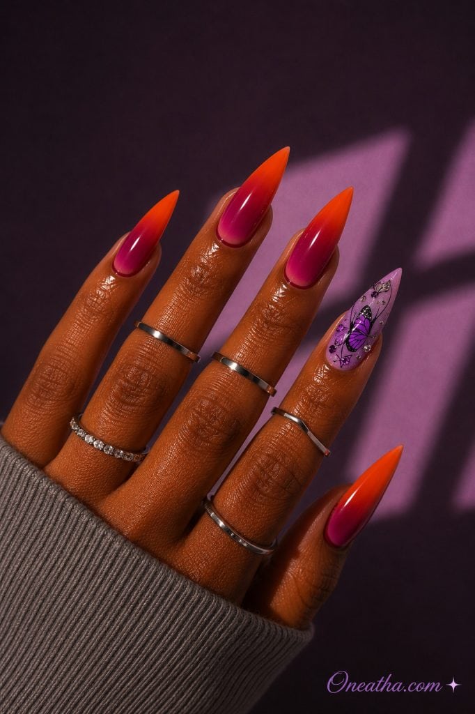 Warm cinnamon-toned hand with sunset ombré stiletto nails and a lilac butterfly accent nail.