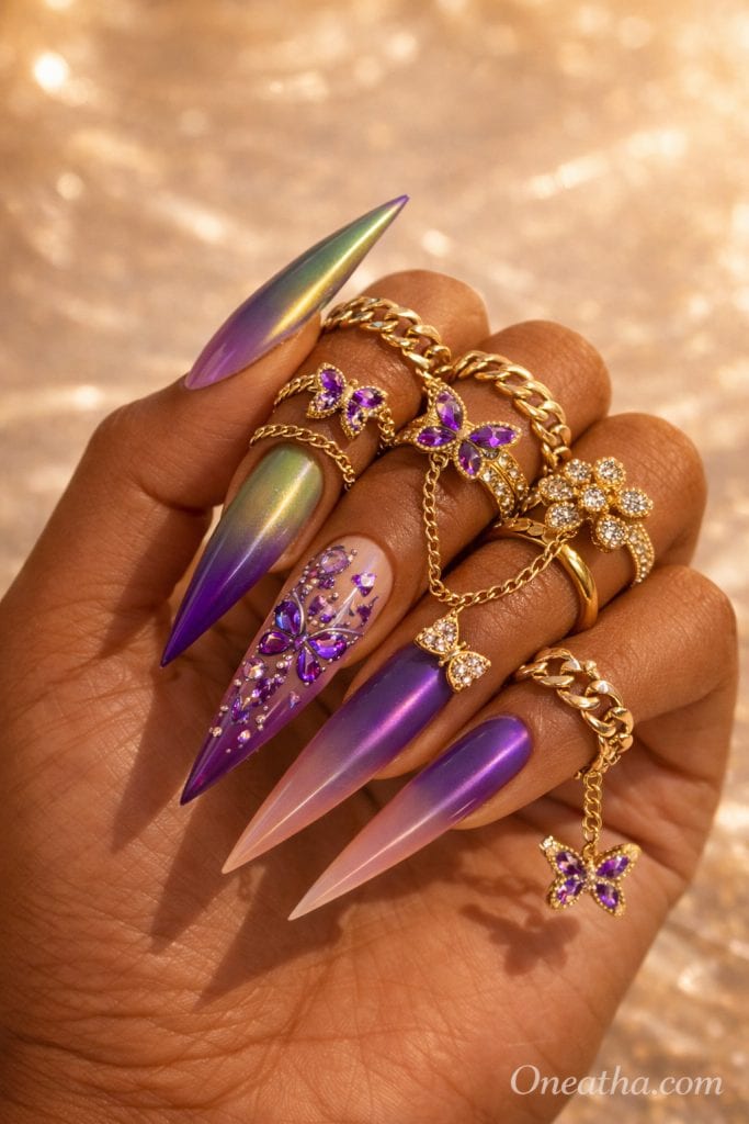 Butterfly Glam luxury nail set by Oneatha featuring long stiletto acrylic nails in purple chrome, lilac ombré, and soft green shimmer tones with hand-placed butterfly crystal designs and gemstone detailing. The look is styled on a warm cinnamon-toned hand wearing gold butterfly chain rings and luxury statement rings under a sunlit glow, showcasing high-end editorial nail art aesthetics by Oneatha.