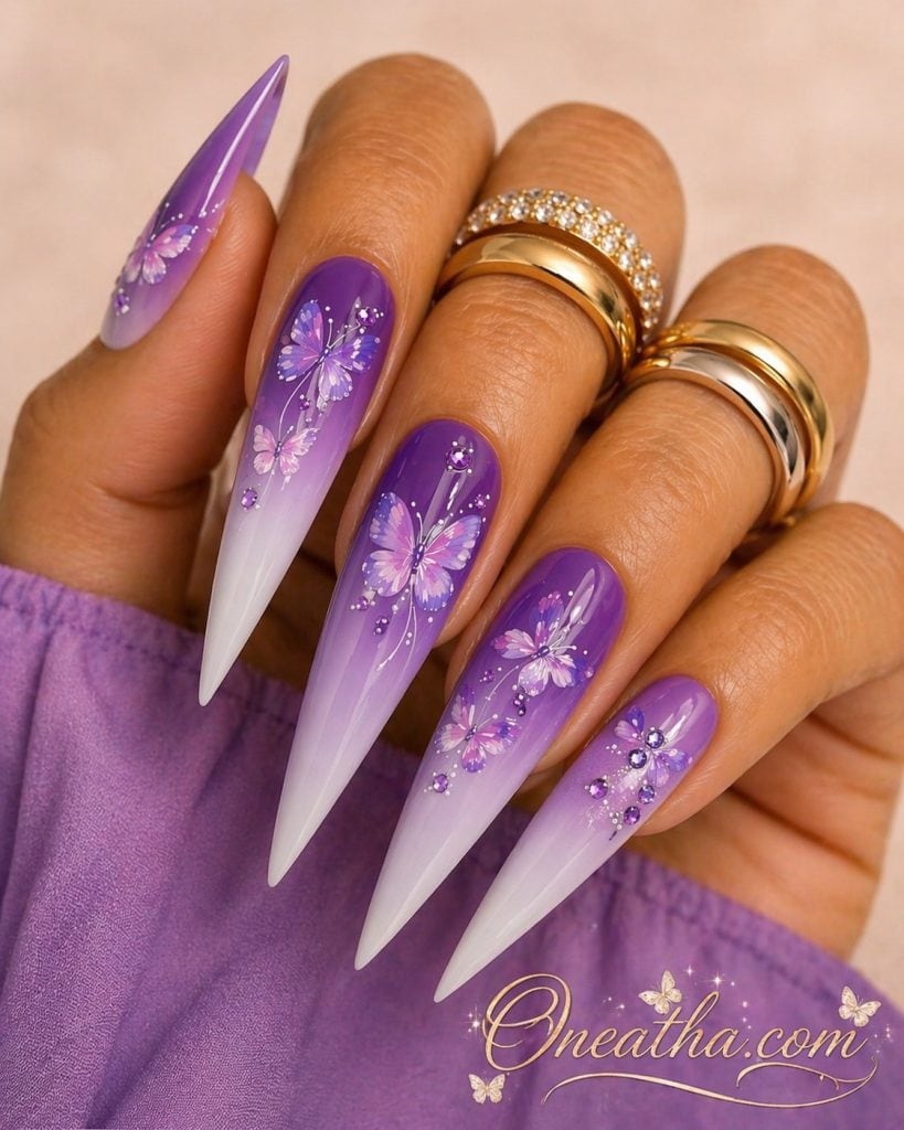 Luxury purple butterfly stiletto nails featuring a soft lilac aura effect that fades into a deep plum purple base. The elegant nail design showcases multiple shades of purple butterfly nail art accented with sparkling purple gemstones. These glamorous stiletto nails offer a high-end, feminine look perfect for summer, special occasions, or year-round wear, pairing beautifully with both casual denim outfits and elegant dresses.