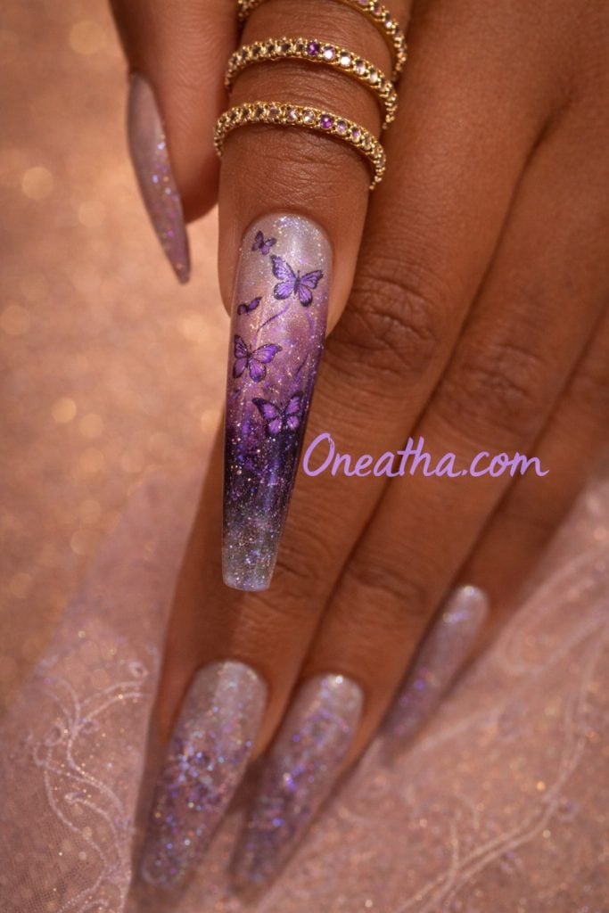 Close-up luxury nail design featuring long coffin-shaped nails with purple ombre shimmer, lilac butterfly detailing, and glitter accents on a warm-toned hand model. Designed by Oneatha, this editorial nail art showcases glam butterfly nail inspiration with chrome sparkle effects and soft gradient artistry. Perfect for luxury nail design inspiration, purple butterfly nails, and aesthetic nail trends. Oneatha.com showcases high-end nail art, beauty inspiration, and signature glam styles for modern luxury looks.