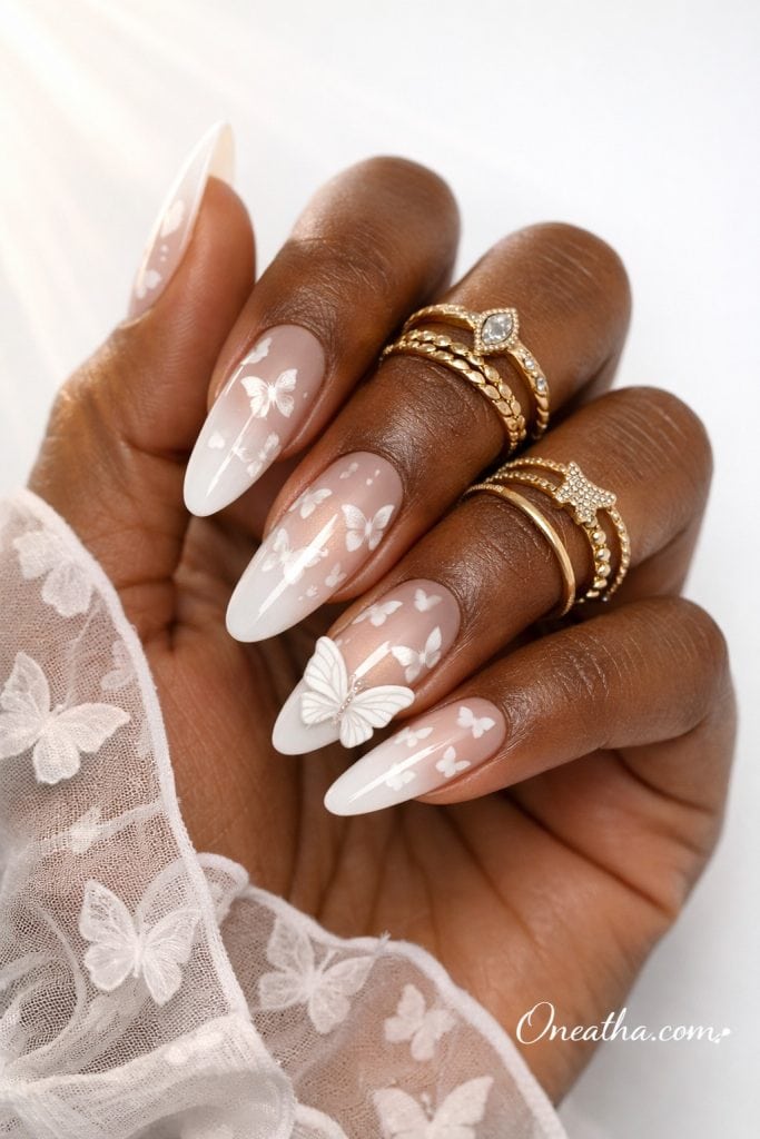 Close-up of a deep cocoa-toned hand featuring long almond-shaped milky white nails with delicate white butterfly designs and a 3D butterfly accent. The nails are styled with glossy French tips, gold rings, and a sheer butterfly blouse against a soft editorial white background.
