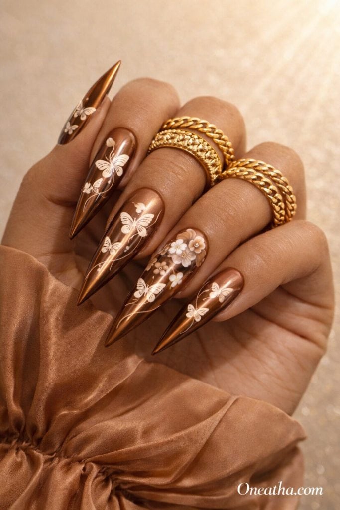 Olive-toned hand with almond-shaped brown chrome nails featuring white butterfly designs and one accent nail with 3D ombré brown and white flowers. The hand is styled with layered gold rings and a silky caramel blouse against a warm beige glowing background.