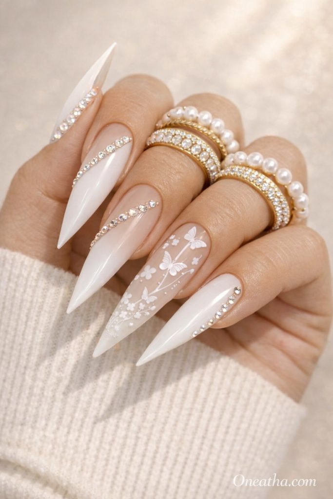 Close-up of a warm beige-toned hand with long stiletto white ombré nails featuring delicate white butterflies, floral accents, and rhinestone curves. The hand is styled with gold and pearl rings against a soft cream sweater and sunlit neutral background.