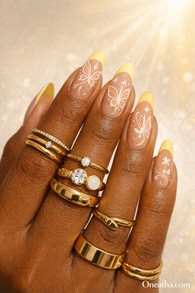 Warm cinnamon-toned hand with almond-shaped nails featuring butter yellow French tips and white butterfly line designs with glossy finish and gold rings. Soft luxury butterfly nail design with a clean editorial aesthetic and golden sunlight background.
