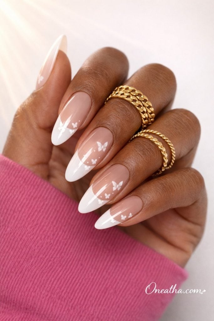 Close-up of a warm cinnamon-toned hand with glossy almond-shaped French tip nails featuring delicate white butterfly designs. Styled with gold rings and a soft pink sweater against a light luxury background.