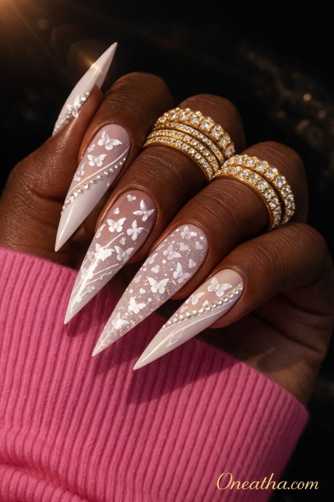 Close-up of a deep cocoa-toned hand with long blush-toned stiletto nails featuring white butterfly designs and gemstone accents. The nails are styled with gold rings and a soft glowing background, creating a romantic luxury aesthetic.
