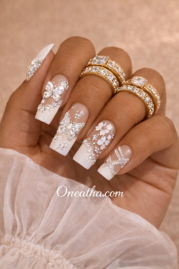 Close-up of a warm cinnamon-toned hand with white French tip nails featuring butterfly designs and sparkling gemstone embellishments. The nails are styled with gold rings and a soft sheer fabric background, creating a bright, elegant luxury look.