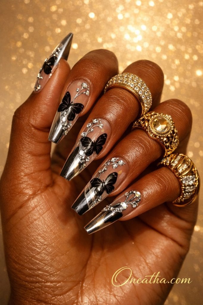 Long coffin nails with black butterfly designs, silver chrome tips, and rhinestone accents on a deep brown hand with gold rings and a glowing background. Luxe black butterfly chrome nail design with clear nude base and sparkling details for a bold, elegant manicure look.