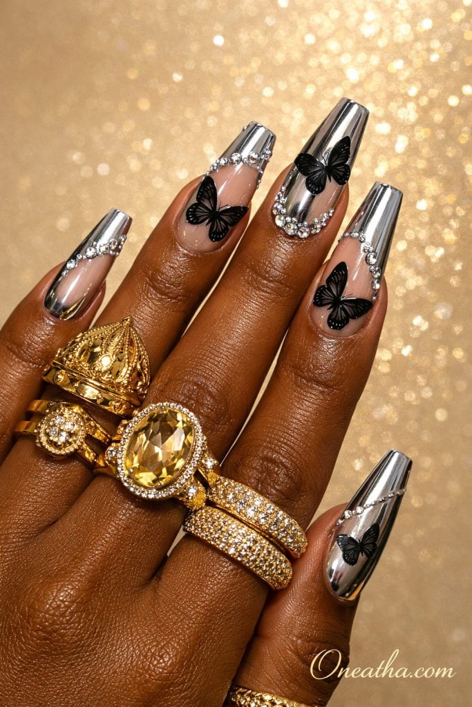 Silver chrome coffin nails with black butterfly designs and rhinestone accents on a deep brown hand with gold rings and a glowing gold background. Luxe black butterfly chrome nail design with reflective finish and crystal details for an elegant, high-shine manicure look.