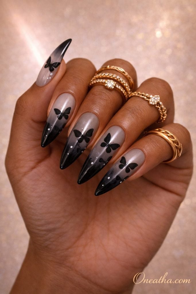 Gray to black ombre almond nails with delicate black butterfly designs on an olive-toned hand with stacked gold rings and a warm glowing background. Glossy butterfly nail design with soft gradient fade and elegant detailing for a modern, luxe manicure look.