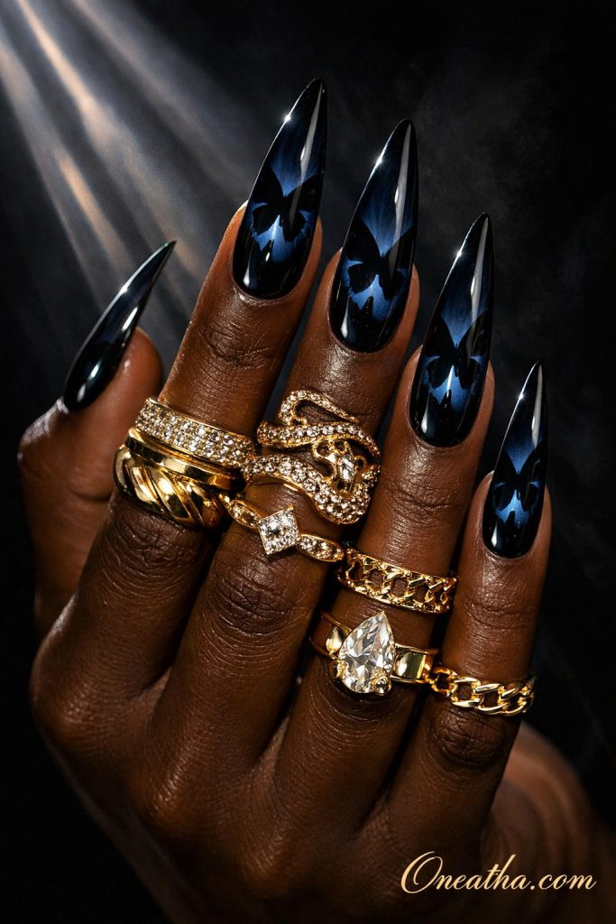 Glossy black stiletto nails featuring subtle butterfly designs on a deep cocoa-toned hand with stacked gold rings and a dramatic dark background. Elegant black butterfly nail design with high shine finish and luxe gold jewelry styling for a bold, sophisticated look.