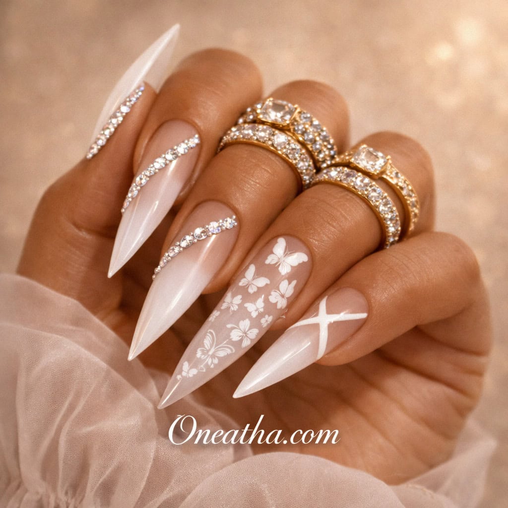 Close-up of a warm cinnamon-toned hand with long milky white stiletto nails featuring delicate butterfly designs and subtle gemstone accents. The nails are styled with gold rings and a soft sheer fabric background, creating a clean and luxurious aesthetic.