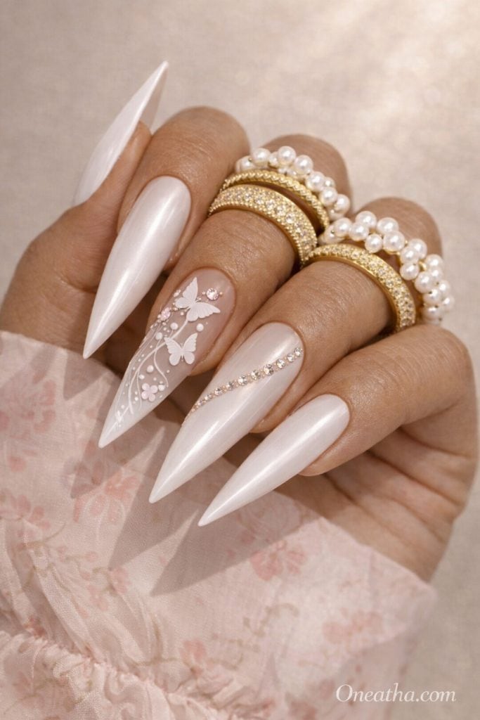 Close-up of a warm cinnamon-toned hand with long coffin white chrome nails featuring butterfly designs, soft ombré detail, and rhinestone accents. Styled with gold and pearl rings against a sheer pink floral blouse with a glowing neutral background.