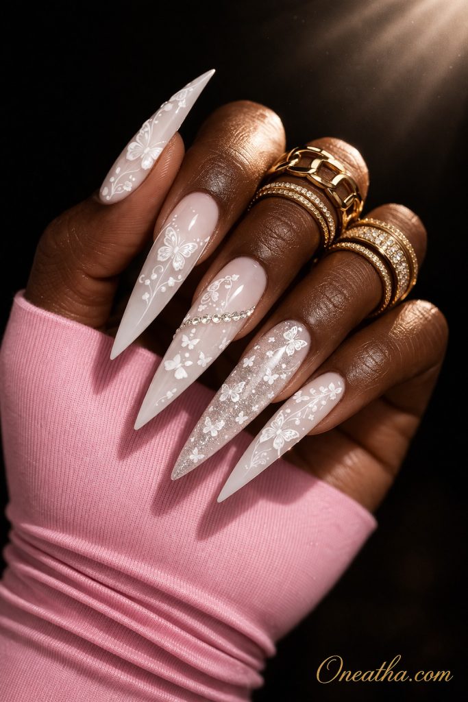 White stiletto nails with delicate butterfly designs and subtle shimmer on a deep brown hand with gold rings and a soft glowing background. Elegant white butterfly nail design with glossy finish and fine detailing for a soft, luxe manicure look.