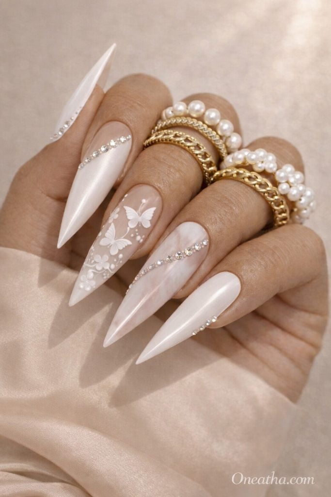 Close-up of a warm tan-toned hand with long stiletto white nails featuring butterfly designs, marble accents, and rhinestone details. Styled with gold and pearl rings against a soft satin neutral background.