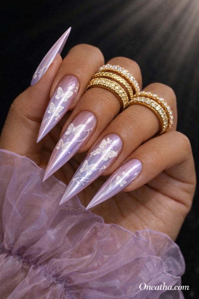 Close-up of a warm-toned hand with long stiletto lavender nails featuring white butterfly designs and a glossy finish. Styled with gold rings and a sheer lilac fabric against a dark luxury background.