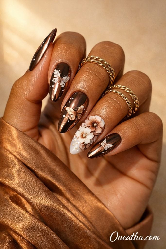 Close-up of a warm cinnamon-toned hand with glossy brown chrome nails featuring white butterfly designs, floral accents, and gemstone details. The nails are styled with gold rings and a silky bronze fabric background, creating a rich, luxe aesthetic.