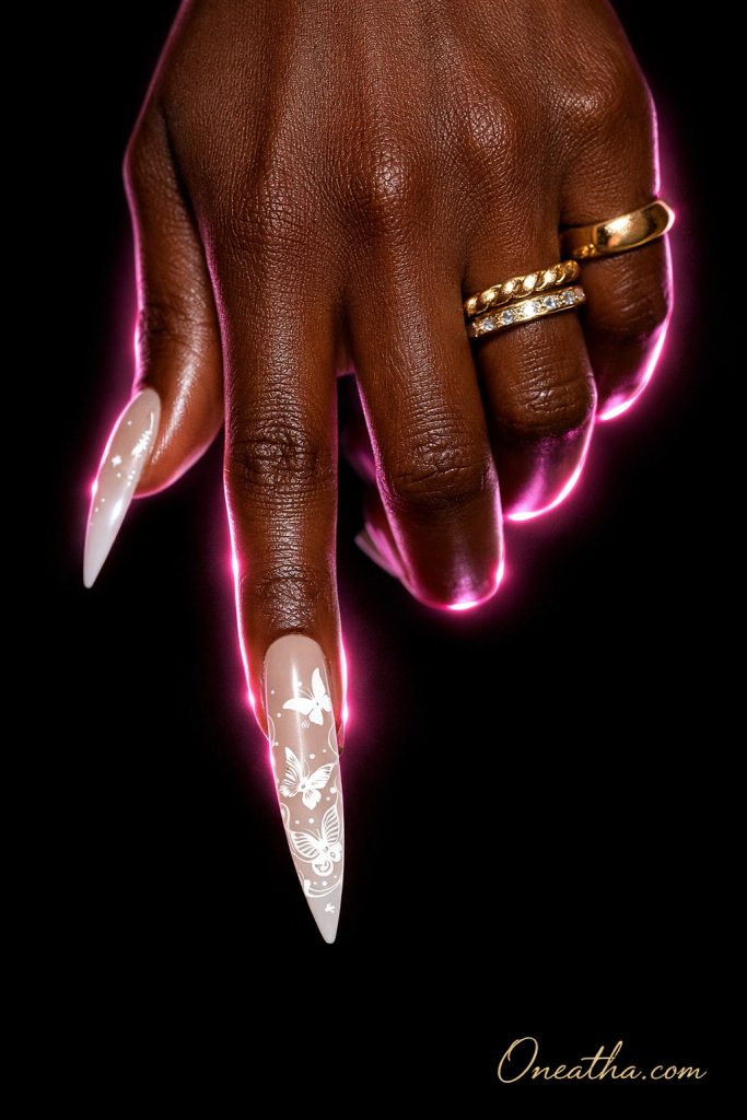 Deep cocoa-toned hand model with long stiletto beige aura nails featuring white butterfly nail art, delicate white line designs, and a glossy pearl finish. Luxury manicure styled with gold rings, pink neon glow lighting, and a black background for a high-end editorial nail aesthetic.