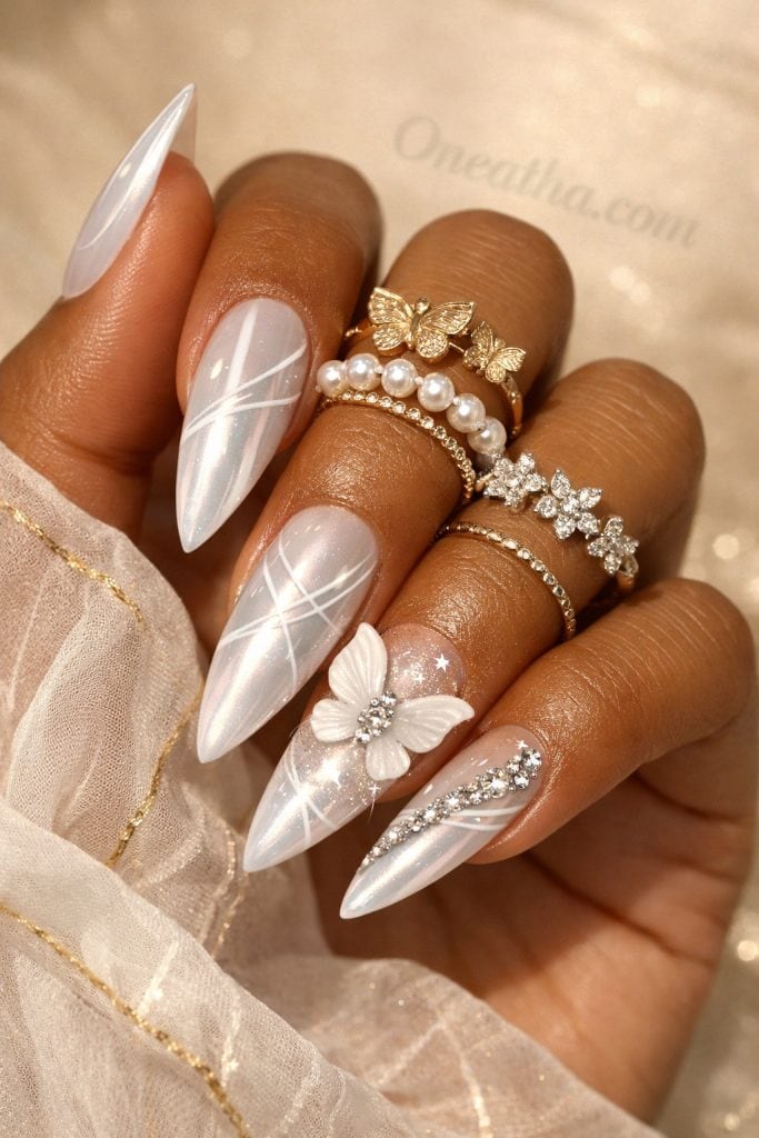Close-up of a warm cinnamon-toned hand with long pearly white nails featuring butterfly designs and sparkling gemstone accents. The nails are styled with gold and pearl rings against a soft, glowing neutral background for a luxe aesthetic.