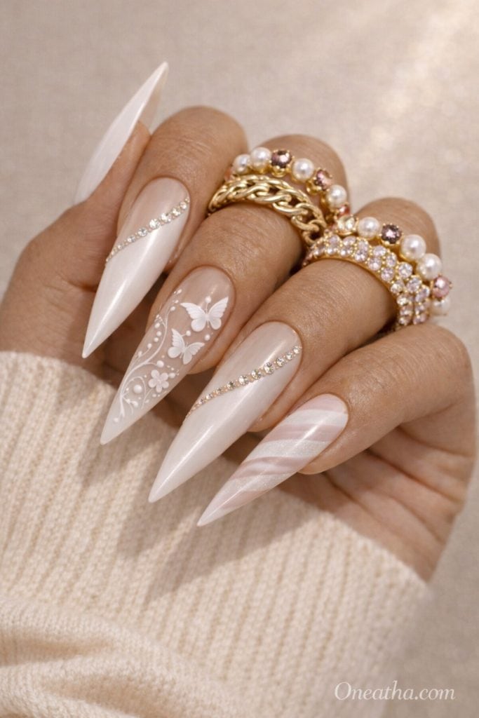 Warm-toned hand with long stiletto milky white nails featuring white butterfly designs, rhinestone accents, and subtle swirl patterns. Styled with stacked gold and pearl rings and a cream sweater against a soft beige luxury background.