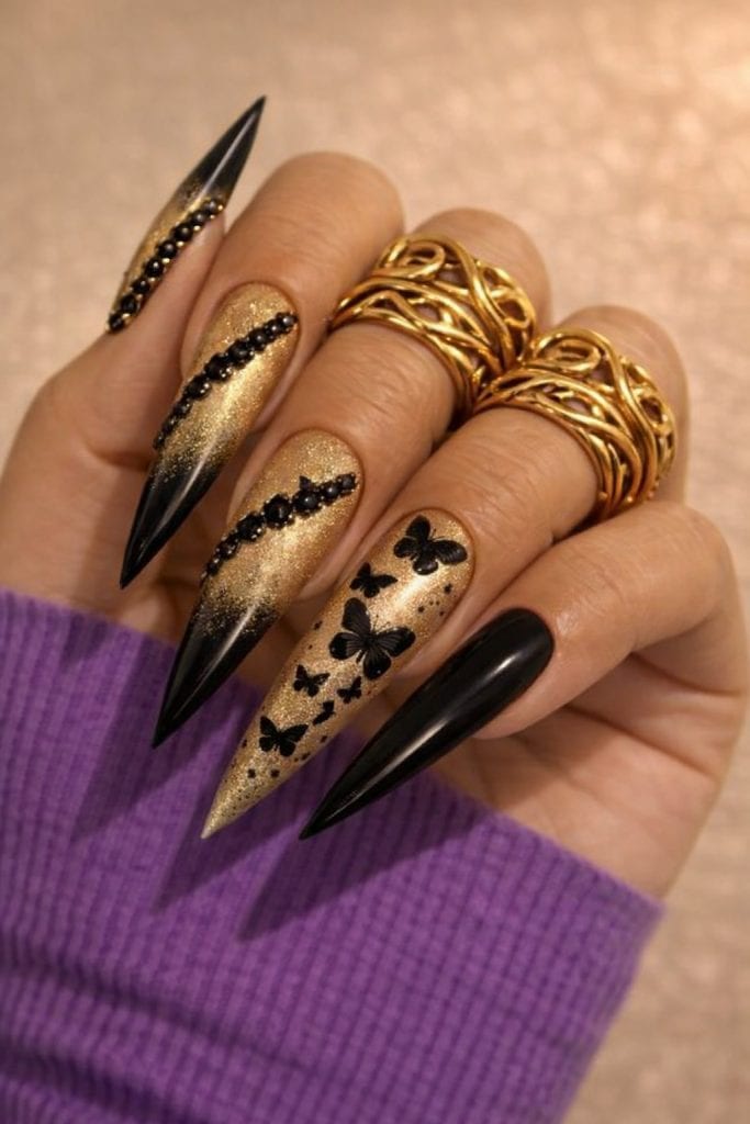 Black and gold stiletto nails with butterfly designs and shimmer gradient on a hand with gold rings and a soft neutral background. Luxe black butterfly nail design with gold sparkle, glossy finish, and bold elegant styling.