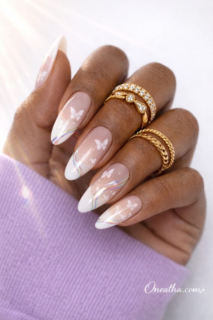 Close-up of an olive-toned hand with almond-shaped ombré white nails featuring delicate butterfly designs and iridescent chrome swirl accents. Styled with gold rings and a lilac sweater against a bright luxury background.