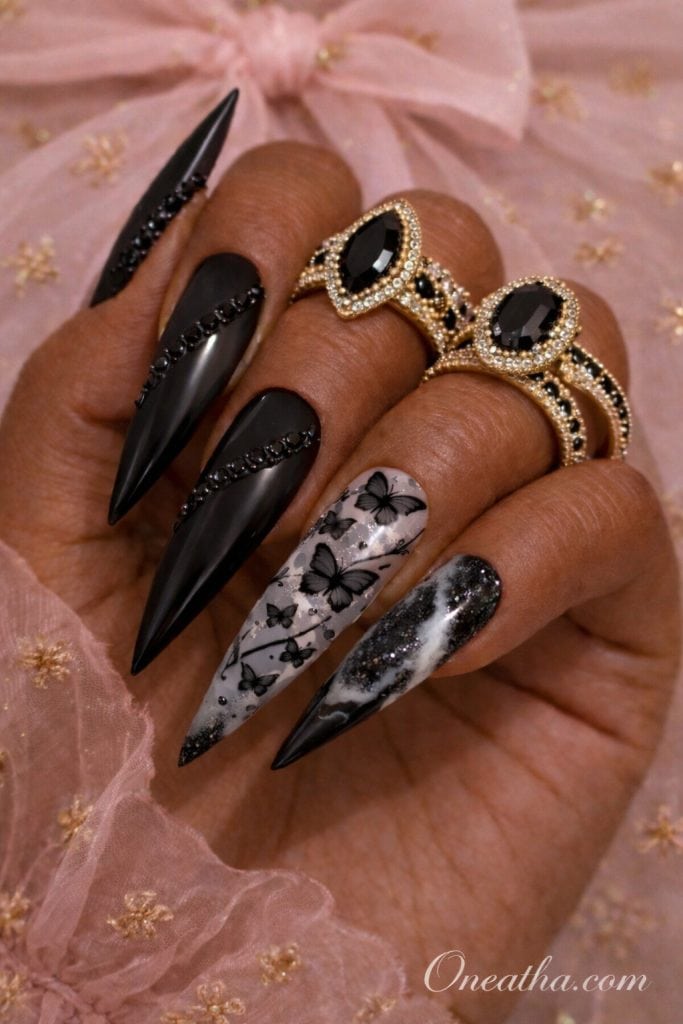 Black stiletto nails with butterfly designs and black-and-white marble accent nails on a hand with gold rings and a soft pink background. Luxe black butterfly nail design with matte and glossy textures for a bold, elegant manicure look.