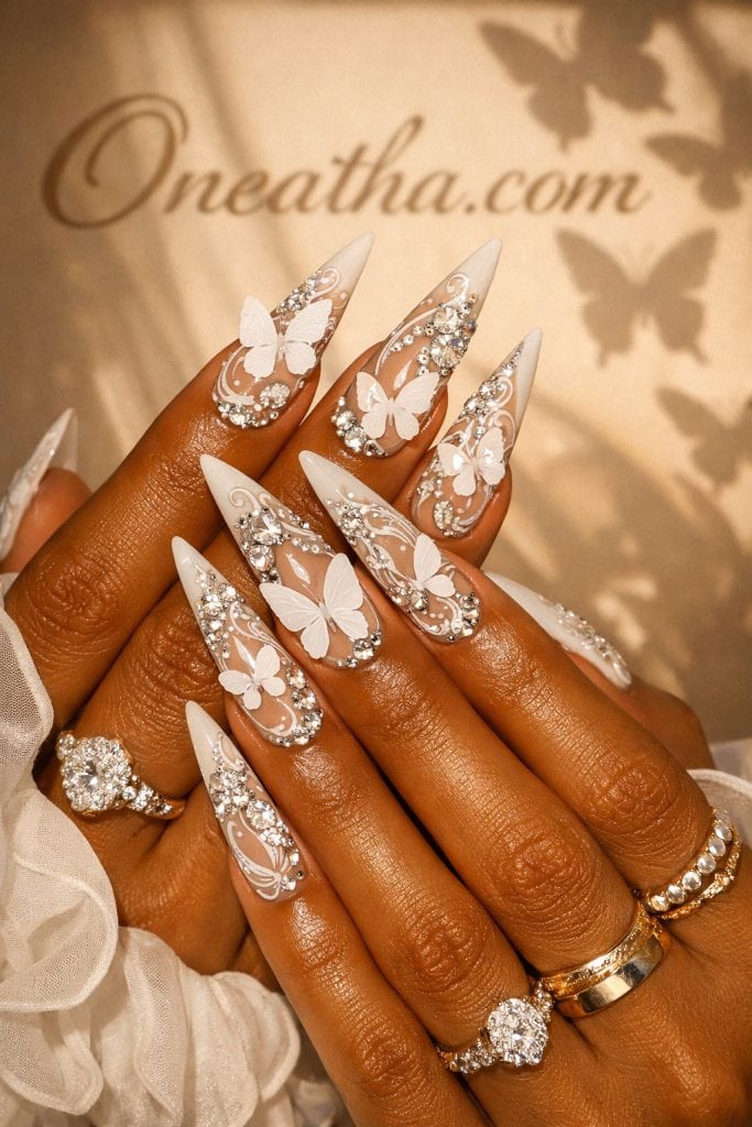 Close-up of a warm deep-toned hand with long stiletto nude and white nails featuring 3D butterfly designs and heavy rhinestone detailing. Styled with gold rings against a glowing beige background