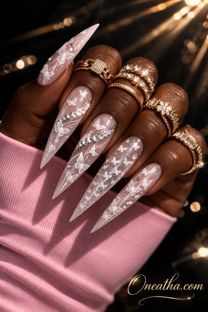 Close-up of a deep cocoa-toned hand with long stiletto nails featuring white butterfly designs and gemstone accents on a glossy nude base. The nails are styled with gold rings and a glowing dark background, creating a bold, luxe aesthetic.