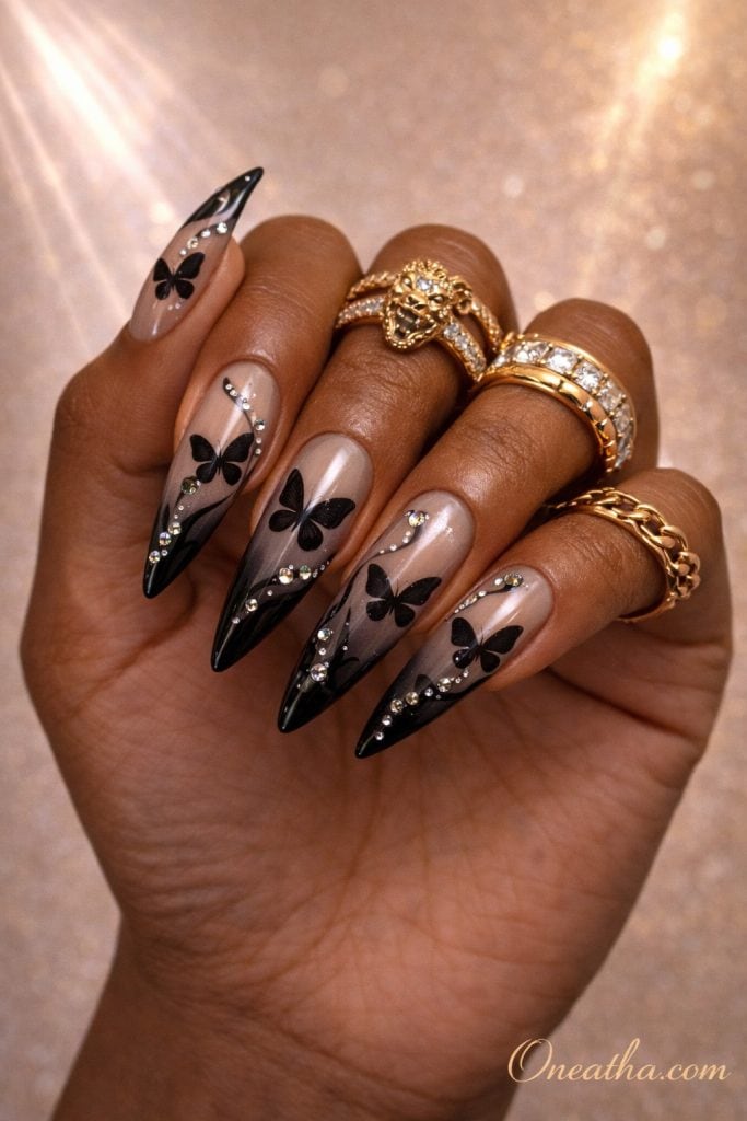 Stiletto nails with black butterfly designs, abstract swirl lines, and rhinestone accents on a rich brown hand with gold rings and a glowing background. Elegant black butterfly nail design with glossy finish and gemstone detailing for a luxe, modern manicure look.