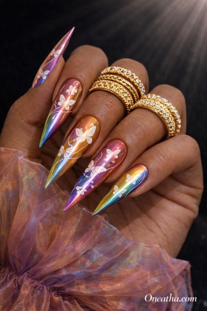 Warm cinnamon-toned hand with long stiletto rainbow chrome nails featuring white butterfly designs and abstract line detailing. Styled with stacked gold rings and a sheer iridescent blouse against a dark luxury background with glowing light beams.