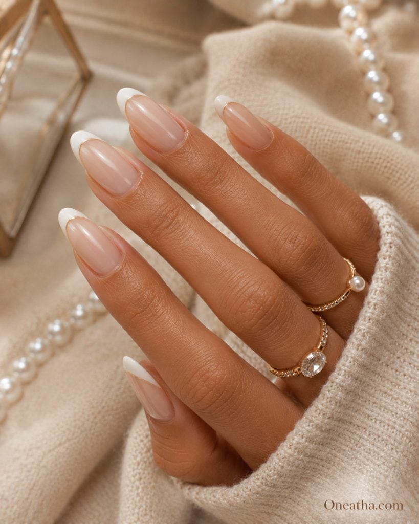 Milky nude French tip almond nails with glossy finish on an elegant hand model, featuring a timeless luxury manicure against a soft neutral background, beauty inspiration by Oneatha.com.
