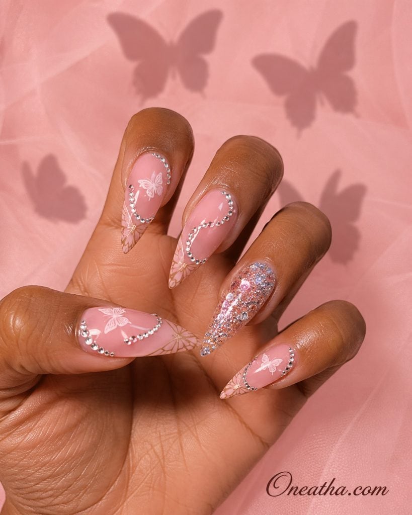 Butterfly nail designs by Oneatha featuring stiletto-shaped nails with blush pink butterfly tips and rhinestone S-shaped accent nail.