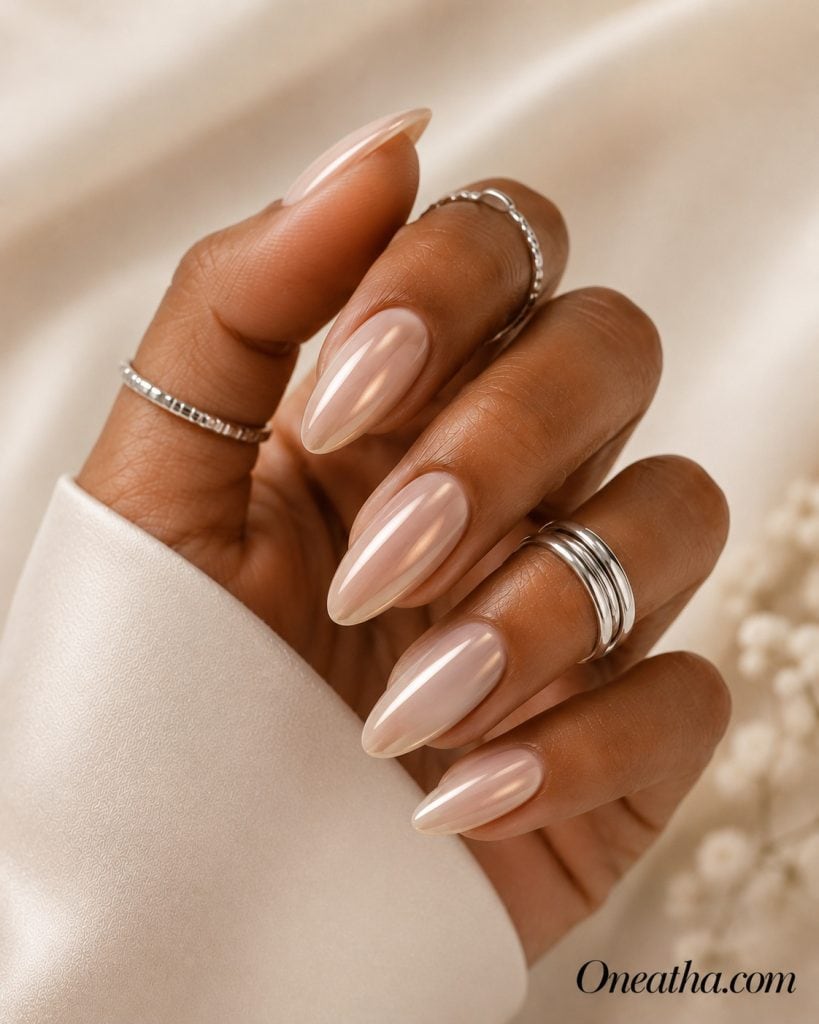 Luxury nail designs by Oneatha featuring translucent nude almond nails with a pearly chrome shimmer, glossy finish, silver luxe rings, and a soft creamy editorial background.
