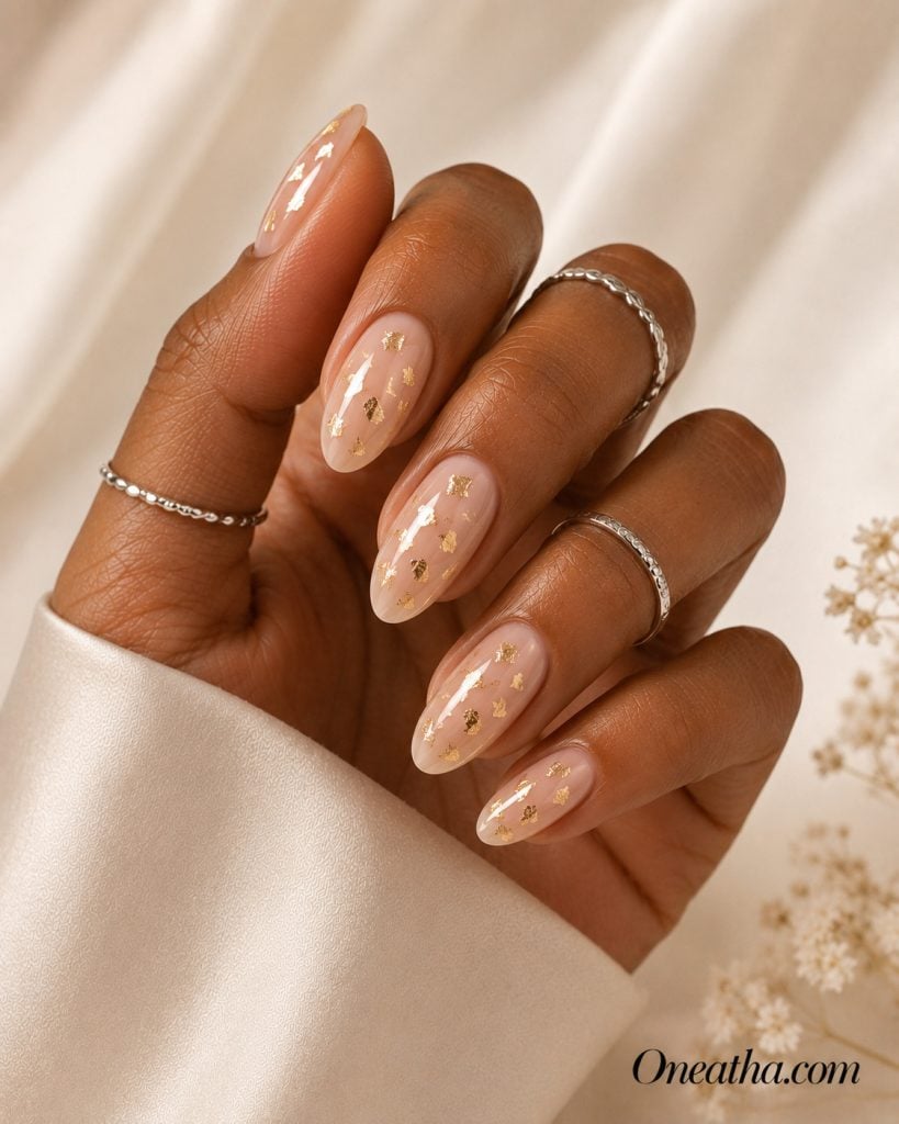 Luxury nail designs by Oneatha featuring sheer nude almond nails with scattered gold foil flakes, a glossy finish, silver rings, and a soft ivory editorial background.