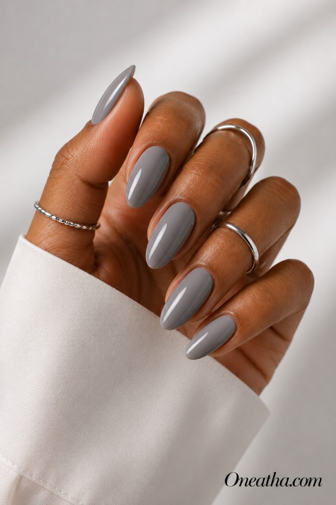 Luxury gray almond nail designs by Oneatha featuring glossy cool gray nails on a warm cinnamon-toned hand with silver rings against a soft white editorial background.