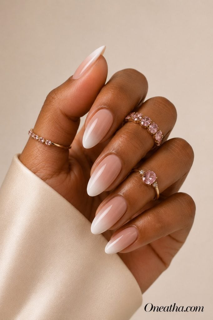 Luxury nail designs by Oneatha featuring glossy nude-to-white ombré almond nails with a seamless gradient, pink gemstone rings, and a soft neutral editorial background.