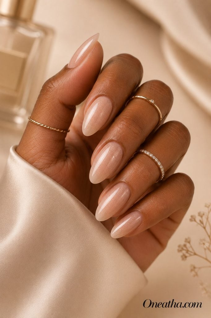 Luxury nail designs by Oneatha featuring sheer nude almond nails with an ultra-fine shimmer overlay, glossy finish, delicate gold rings, and a soft beige editorial background.