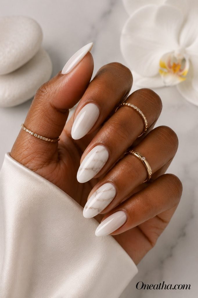 Luxury nail designs by Oneatha featuring glossy milky white almond nails with soft marble accent nails, gold rings, and a clean gray-white editorial spa-inspired background.