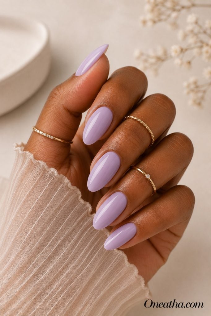 Luxury nail designs by Oneatha featuring glossy lavender almond nails, gold rings, and a soft neutral editorial background with a pastel, elegant aesthetic.