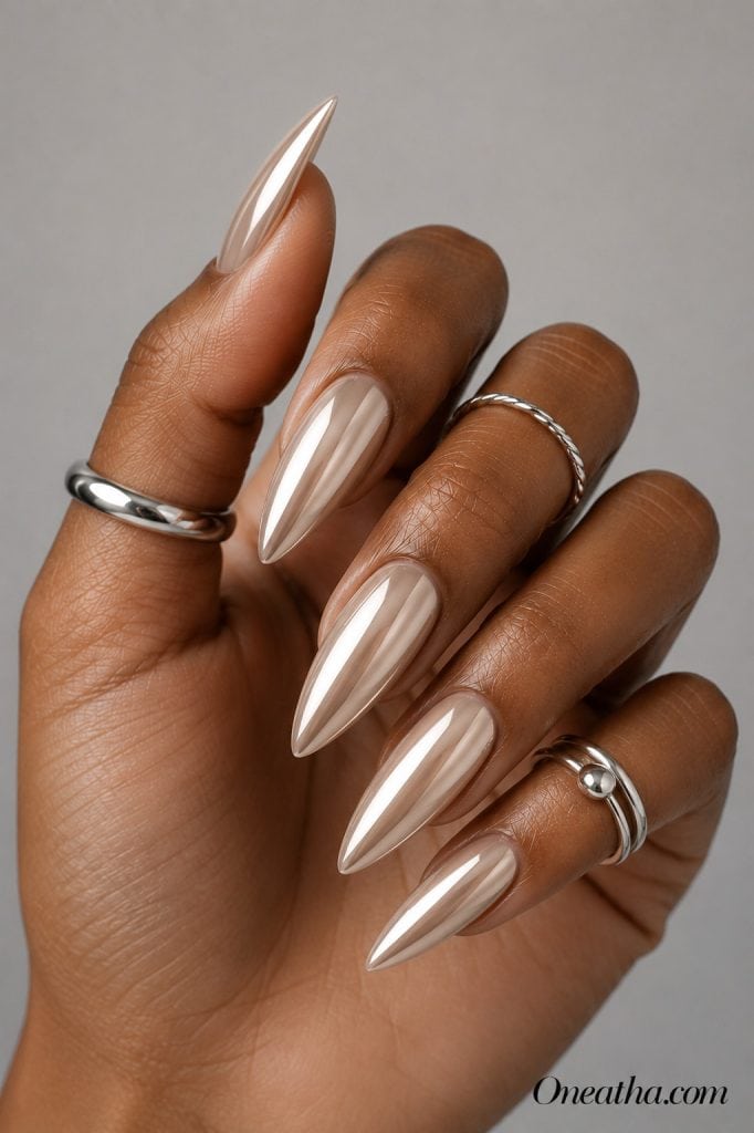 Luxury nail designs by Oneatha featuring light beige chrome almond nails with a reflective mirror finish, silver rings, and a neutral gray editorial background.